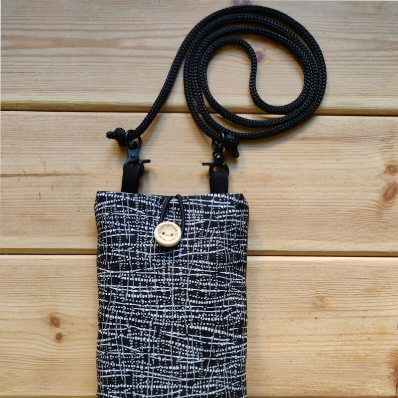 Pode incluir: Uma bolsa para telefone com padr&atilde;o preto e branco com uma al&ccedil;a ajust&aacute;vel preta. A bolsa tem um pequeno fecho de bot&atilde;o de madeira.