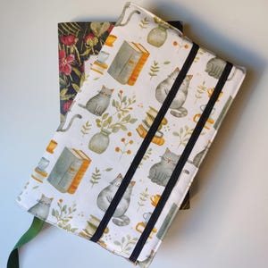 May include: A book cover with a white background featuring a pattern of grey cats, books, vases, and foliage. The cover has black elastic bands and a green ribbon bookmark. Another book cover with a floral pattern is visible behind it.