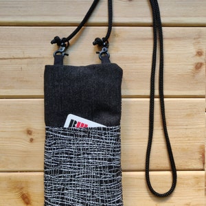Pode incluir: Uma bolsa de ombro de denim preta com um bolso frontal estampado em branco e preto. A bolsa tem uma al&ccedil;a ajust&aacute;vel preta e um pequeno cart&atilde;o vermelho &eacute; vis&iacute;vel no bolso.