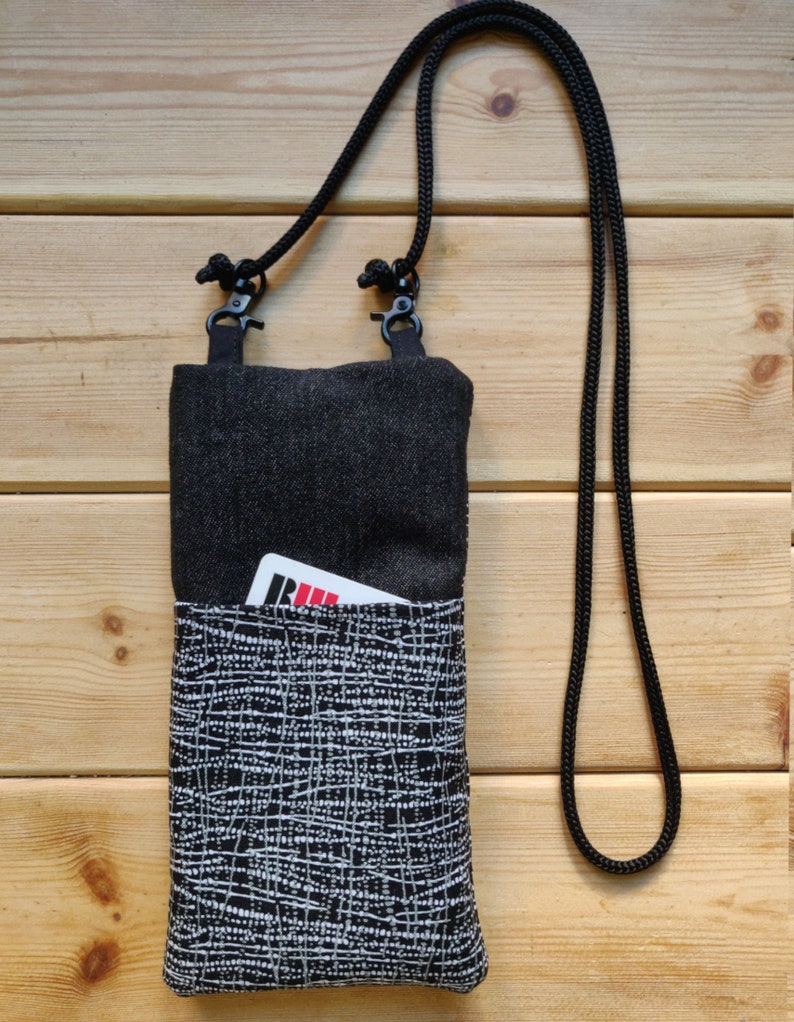 Pode incluir: Uma bolsa de ombro de denim preta com um bolso frontal estampado em branco e preto. A bolsa tem uma al&ccedil;a ajust&aacute;vel preta e um pequeno cart&atilde;o vermelho &eacute; vis&iacute;vel no bolso.