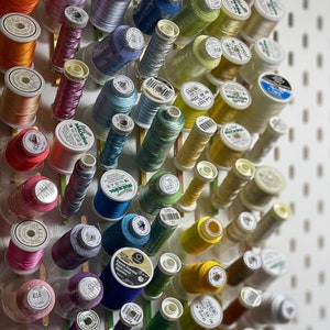 May include: A white pegboard organizer filled with spools of thread in various colors, including red, orange, pink, purple, blue, green, yellow, and black. The spools are arranged in rows, creating a visually appealing display for sewing supplies.