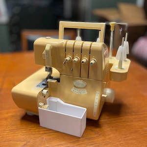 May include: A beige Eclipse Special Edition serger sewing machine. The machine has multiple dials, thread guides, and a white waste bin. The machine is on a wooden surface.