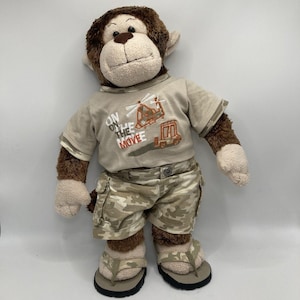 May include: A plush monkey toy with brown and cream fur wearing a beige t-shirt with a helicopter and truck graphic and the text ON THE MOVE, camouflage cargo shorts, and beige flip-flops. The monkey has a stitched nose and mouth.