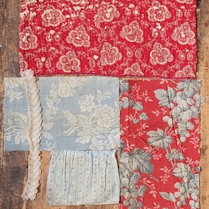 May include: Assortment of vintage fabric swatches in various floral patterns. Includes a quilted red piece with white floral designs, light blue fabric with white flowers, and a red fabric with gray leaves and grapes. A piece of lace is also present.
