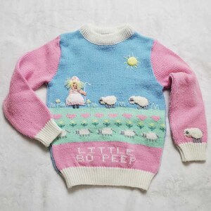 May include: A colorful Little Bo Peep sweater with a sky blue upper section, pink sleeves, and a pink and white lower section. The sweater features embroidered sheep, a sun, and the words "Little Bo Peep".