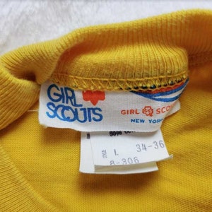 May include: Close-up of a yellow Girl Scouts t-shirt collar with a tag. The tag reads "GIRL SCOUTS NEW YORK" in blue text with a red heart logo. The shirt size is L, with measurements 34-36.