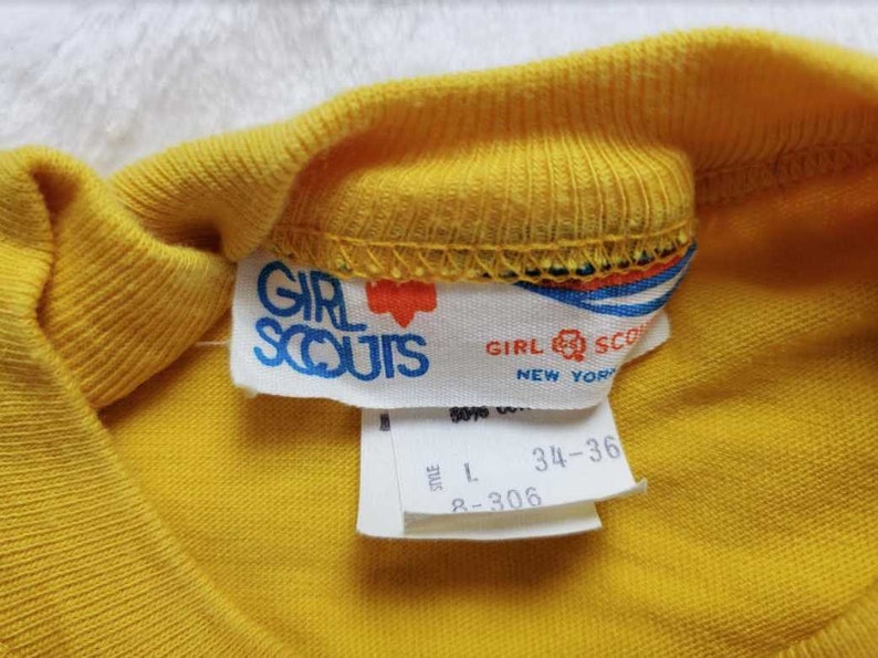 May include: Close-up of a yellow Girl Scouts t-shirt collar with a tag. The tag reads "GIRL SCOUTS NEW YORK" in blue text with a red heart logo. The shirt size is L, with measurements 34-36.