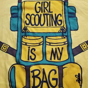May include: Yellow t-shirt with a teal and yellow backpack graphic. The backpack design includes the text "GIRL SCOUTING IS MY BAG" in black lettering. The shirt is made of a soft material.