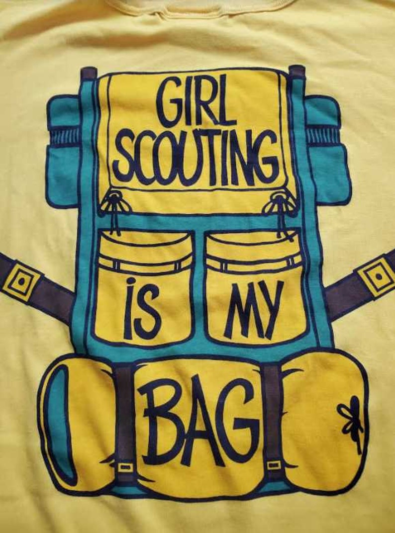 May include: Yellow t-shirt with a teal and yellow backpack graphic. The backpack design includes the text "GIRL SCOUTING IS MY BAG" in black lettering. The shirt is made of a soft material.