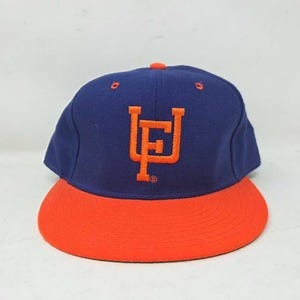 May include: A navy blue baseball cap with an orange peak. The cap has an orange embroidered logo on the front. The cap has a structured crown and a flat peak. A classic style, suitable for casual wear.
