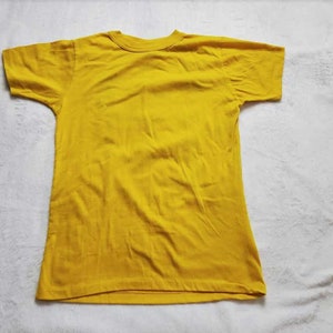 May include: A solid yellow t-shirt with a crew neck and short sleeves. The shirt is made of a soft fabric and is laid flat on a white surface. The t-shirt is a basic, casual garment.