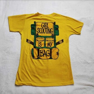 May include: A yellow t-shirt with a graphic of a green and brown backpack. The backpack design has the text "GIRL SCOUTING IS MY BAG" in black letters. The shirt is made of a soft material.