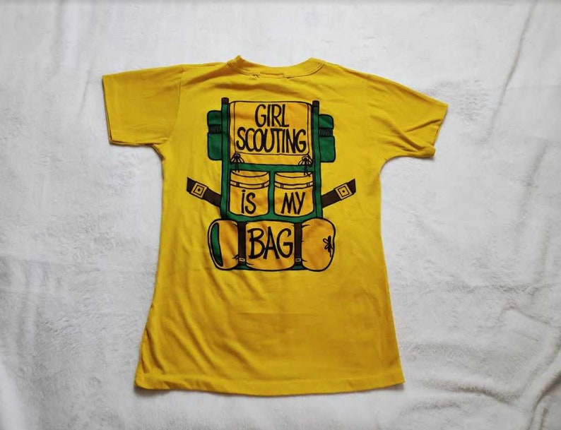 May include: A yellow t-shirt with a graphic of a green and brown backpack. The backpack design has the text "GIRL SCOUTING IS MY BAG" in black letters. The shirt is made of a soft material.