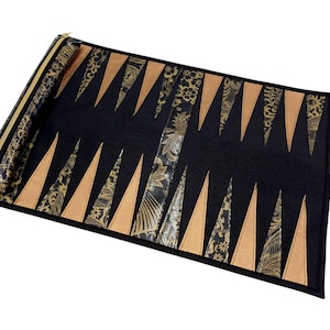 May include: A black and gold backgammon board with a rolled-up section and a strap. The board features alternating tan and black triangles, with gold floral patterned fabric accents. The zipper and trim are gold.