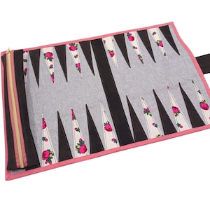 Op de afbeelding: Een backgammonbord in een oprolbare stijl. Het bord heeft een grijze speeloppervlakte met zwarte en witte driehoeken. De witte driehoeken zijn versierd met roze rozen. Het heeft een roze rand en een gouden rits.