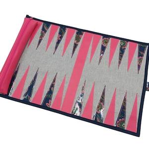 May include: A backgammon board in a roll-up design. The board features a gray playing surface with pink and navy blue accents. The pink elements form the points, and the navy blue is used for the border and the floral patterned points. A pink zippered pouch is attached.