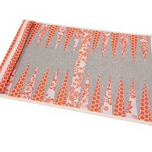 May include: A fabric backgammon board in a rolled-up position. The board features a gray playing surface with orange and white triangular points. The border and zipper are orange with white polka dots. A beige strap is attached.