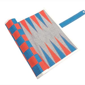 Puede incluir: Un tablero de backgammon enrollado con una correa azul. El tablero presenta un campo central gris con triángulos rojos y azules alternos. Los bordes tienen un patrón de tablero de ajedrez rojo y azul.