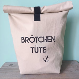 Op de afbeelding: Een beige canvas draagtas met een zwarte band en de tekst "Brötchen Tüte" en een anker dat op de voorkant is gedrukt.