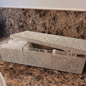 May include: A clear plastic box with a lid covered in rhinestones. The box is divided into compartments and is designed to hold cotton rounds or other small items.