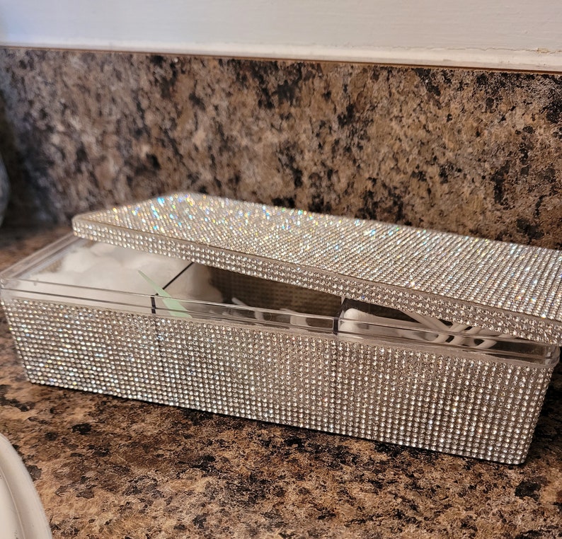 May include: A clear plastic box with a lid covered in rhinestones. The box is divided into compartments and is designed to hold cotton rounds or other small items.