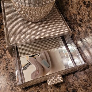 May include: A two-tiered mirrored tray with a clear acrylic drawer. The tray is covered in rhinestones and has a mirrored surface. The drawer is also covered in rhinestones and has a clear acrylic handle.