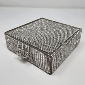 May include: A small, square-shaped box covered in sparkling rhinestones. The box has a small drawer on the front.