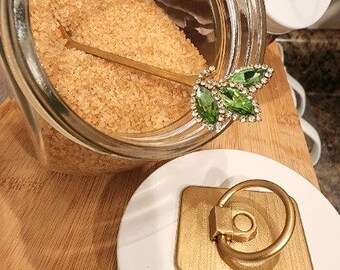May include: A glass jar with a gold lid and a gold spoon inside. The jar is filled with brown sugar. The jar is sitting on a wooden surface. There is a white ceramic lid with a gold ring handle next to the jar.