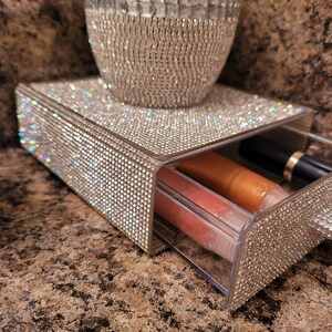 May include: A clear plastic drawer with two lip glosses inside. The drawer is decorated with rhinestones and sits on top of a larger rhinestone-covered container.