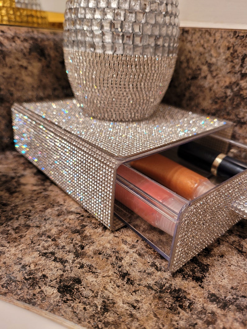 May include: A clear plastic drawer with two lip glosses inside. The drawer is decorated with rhinestones and sits on top of a larger rhinestone-covered container.
