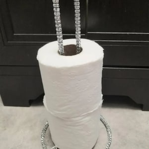 May include: A silver paper towel holder with a rhinestone-encrusted handle and base. The holder is holding a roll of white paper towels.
