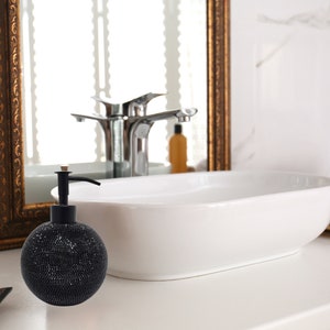 May include: A black and silver soap dispenser with a round, textured bottle and a pump top. The dispenser is sitting on a white countertop in a bathroom.