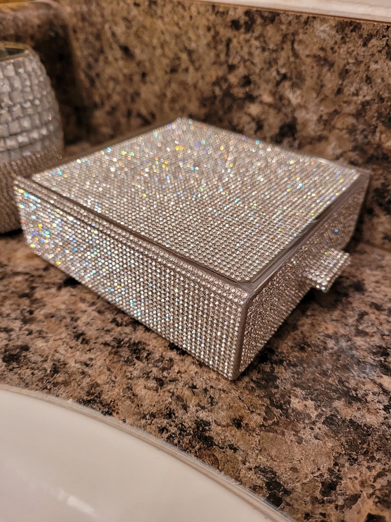 May include: A small, square, silver-colored box covered in clear rhinestones. The box has a hinged lid and a small pull handle on the side.