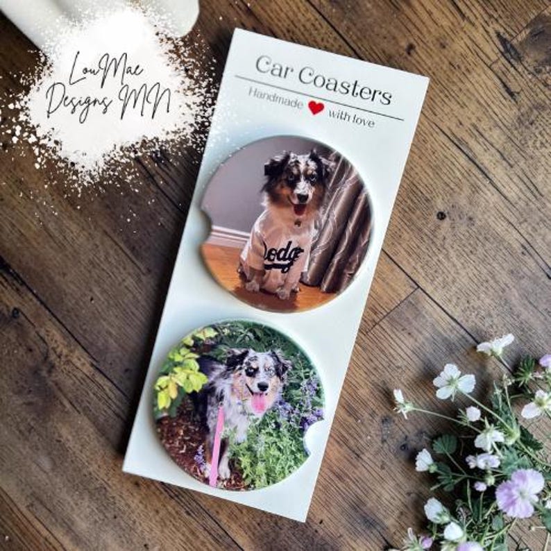 May include: Two car coasters with dog photos. One coaster shows a dog wearing a shirt that says "dodge". The other coaster shows a dog with its tongue out in a garden. The coasters are on a white card that says "Car Coasters" and "Handmade with love".