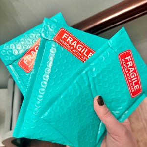 May include: Three teal bubble mailers with red "FRAGILE HANDLE WITH CARE" stickers. The mailers are held in a hand, showcasing their textured surface. The image suggests a focus on packaging and shipping.