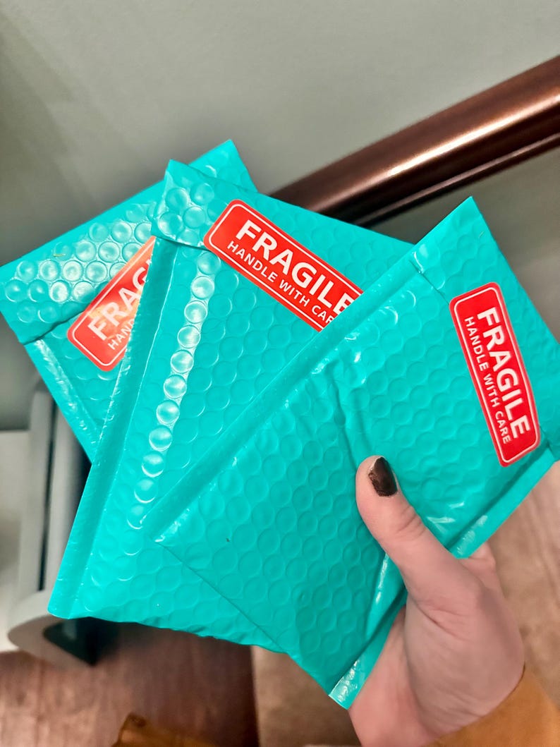May include: Three teal bubble mailers with red "FRAGILE HANDLE WITH CARE" stickers. The mailers are held in a hand, showcasing their textured surface. The image suggests a focus on packaging and shipping.