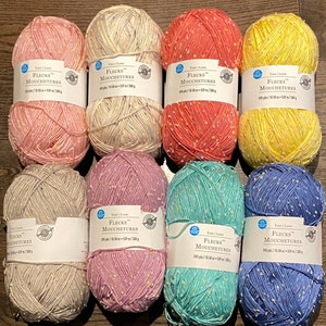 May include: Eight balls of yarn in a variety of pastel colours, including pink, yellow, blue, and green. Each ball has a white label with the text "Flecks Mouchetures 510 yds/10.58 oz - 539 m/300 g" printed on it.
