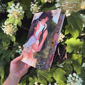 May include: A book with a romantic illustration of a couple embracing, with a woman in a white dress and a man in a green coat. The book's edges feature floral and decorative designs. The book is held in front of a background of green leaves and white flowers.