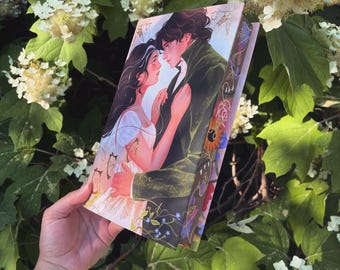 May include: A book with a romantic illustration of a couple embracing, with a woman in a white dress and a man in a green coat. The book's edges feature floral and decorative designs. The book is held in front of a background of green leaves and white flowers.