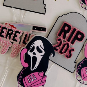 May include: A collection of acrylic charms with a horror theme. Designs include a tombstone with "RIP 20s" in pink glitter, a ghost face with a pink telephone, and a knife. The charms are on a white background.