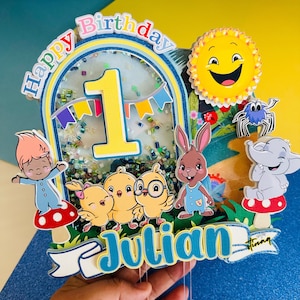 May include: A colorful Happy Birthday cake topper with a yellow number one, a blue bunny, three yellow chicks, a blue gnome, a purple spider, a gray elephant, and a yellow sun. The topper also includes a blue banner with the name Julian.