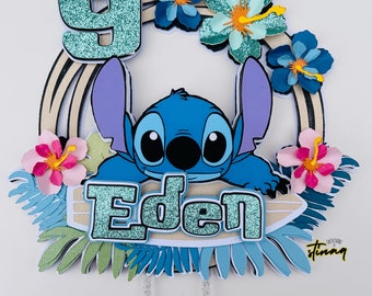 Op de afbeelding: Een blauwe en turquoise taarttopper met het personage Stitch, het cijfer 9 en de naam "Eden". Versierd met tropische bloemen en bladeren, ideaal voor een verjaardagsfeest.