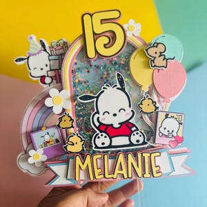 May include: A pink and blue cake topper with a white dog character, a rainbow, and the number 15. The topper also includes the name "Melanie" in yellow letters.