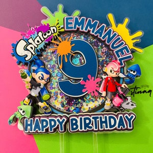 May include: A colorful cake topper featuring the word "Splatoon" and the name "Emmanuel" above a large number 9. The words "Happy Birthday" are at the bottom. The topper includes character figures and ink splatters.