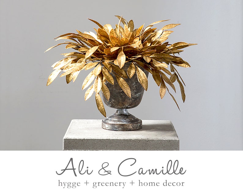 May include: Gold leaf branches in a silver metal urn on a gray concrete pedestal. The urn has a classic design with a round base and a flared top.