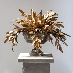 May include: A gold metal planter with a gold leafy plant on a gray pedestal. The planter has a round bowl shape with a pedestal base.
