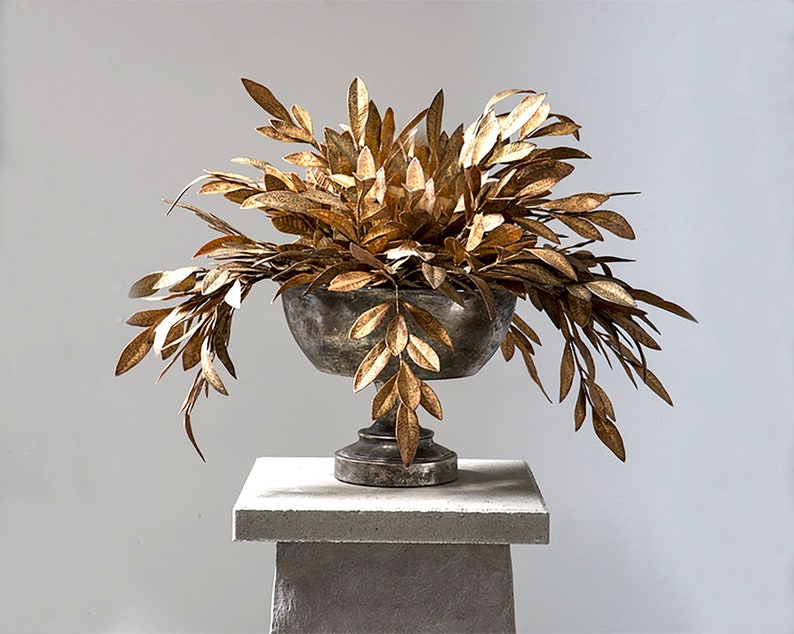 May include: A gold metal planter with a gold leafy plant on a gray pedestal. The planter has a round bowl shape with a pedestal base.