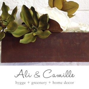 May include: A rustic brown wooden box planter filled with white and green artificial flowers. The text "Ali & Camille" and "hygge + greenery + home decor" is printed below the planter.