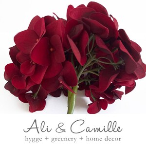 May include: A cluster of deep red artificial hydrangea flowers with green stems. The flowers have a velvety texture and are arranged in a dense, rounded shape. The image includes the text "Ali & Camille hygge + greenery + home decor".