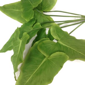 May include: A cluster of artificial green leaves with prominent veining. The leaves are heart-shaped and arranged on slender green stems, bound together with a brown tie. The background is plain white.